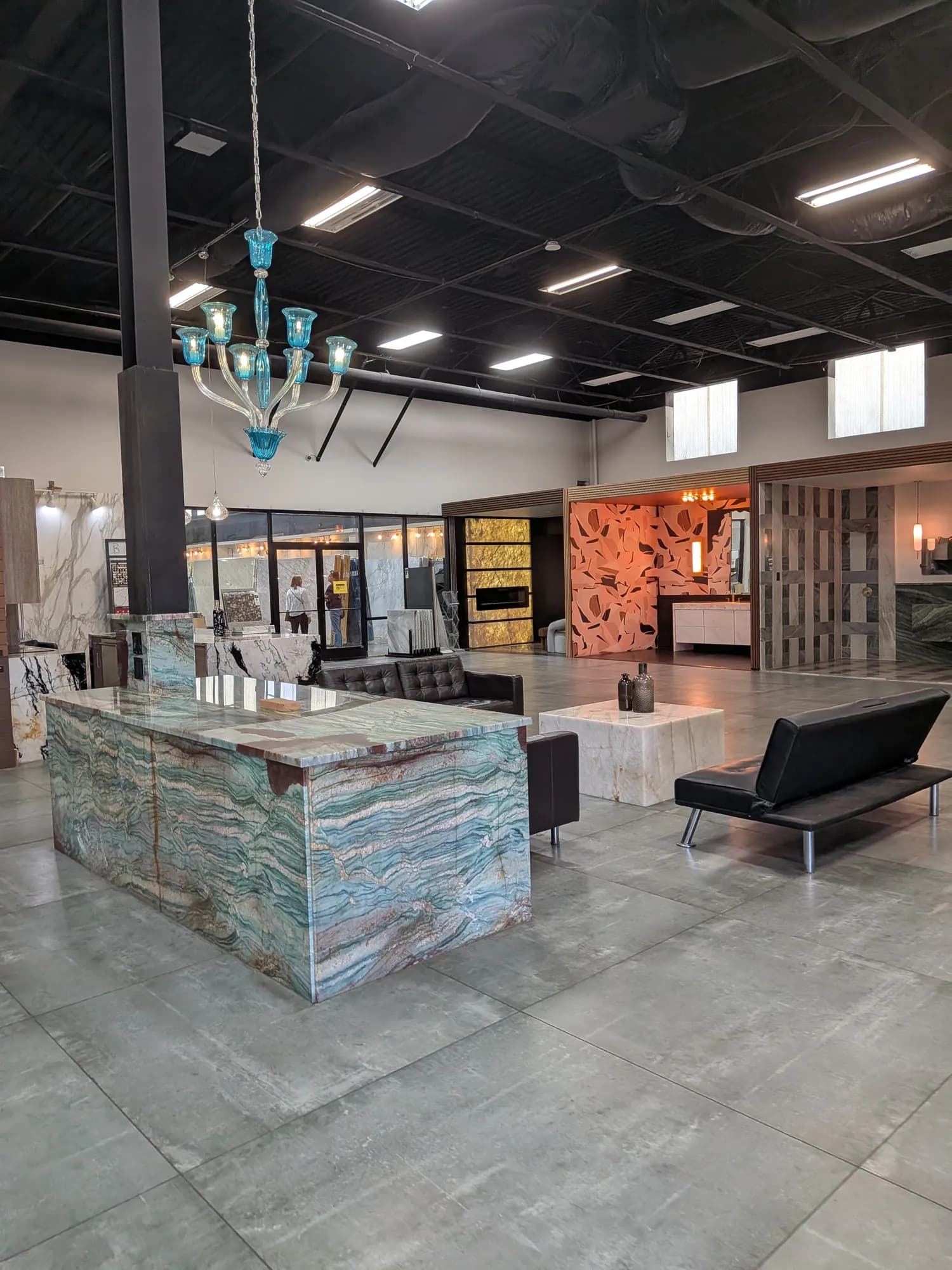Showroom floor with a waterfall-edge island in blue-green banded stone, a turquoise glass chandelier and a backlit stone wall beyond