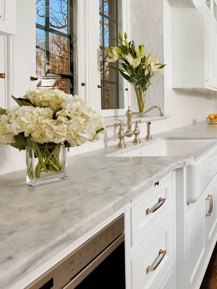 A kitchen perimeter run in soft grey marble with an apron sink, beneath a window