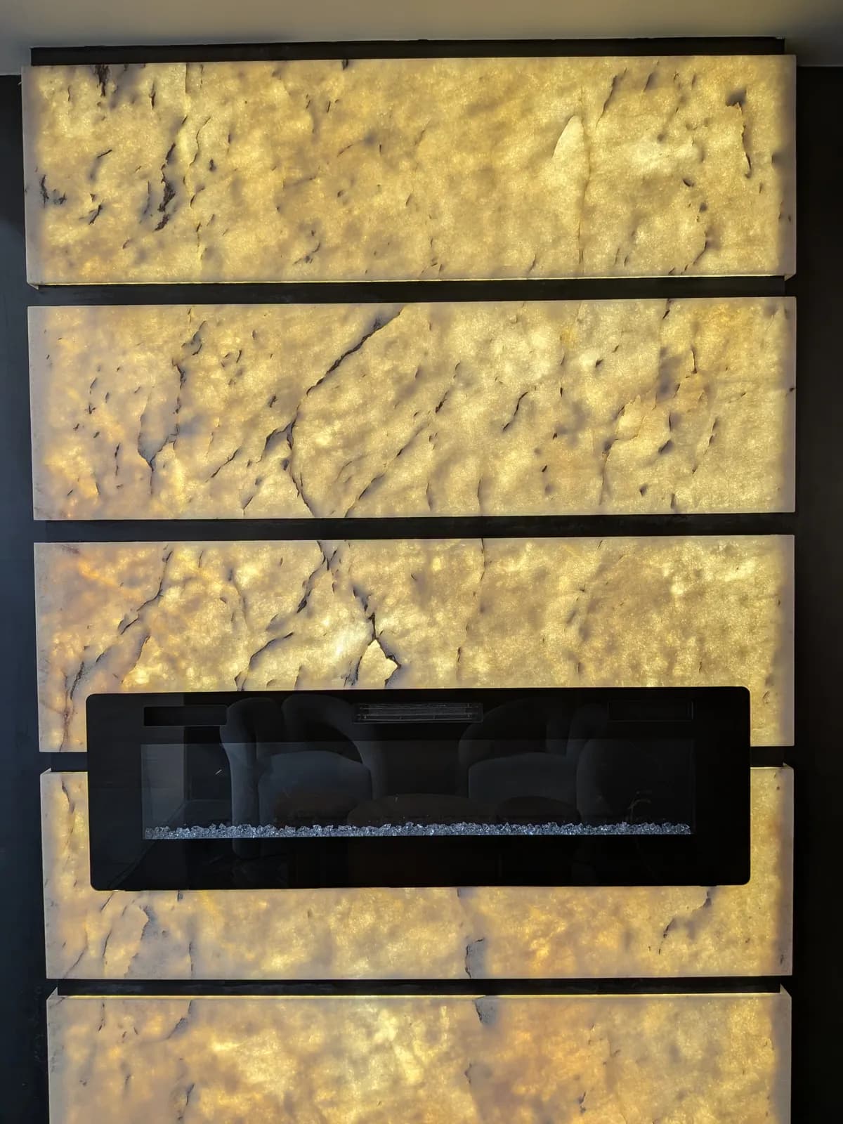 A wall of backlit golden stone panels around a black linear fireplace