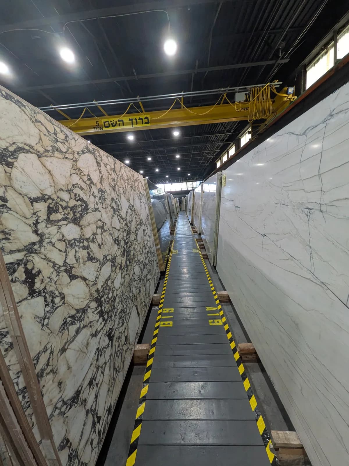 A narrow aisle between tall polished slabs, with a yellow overhead crane above